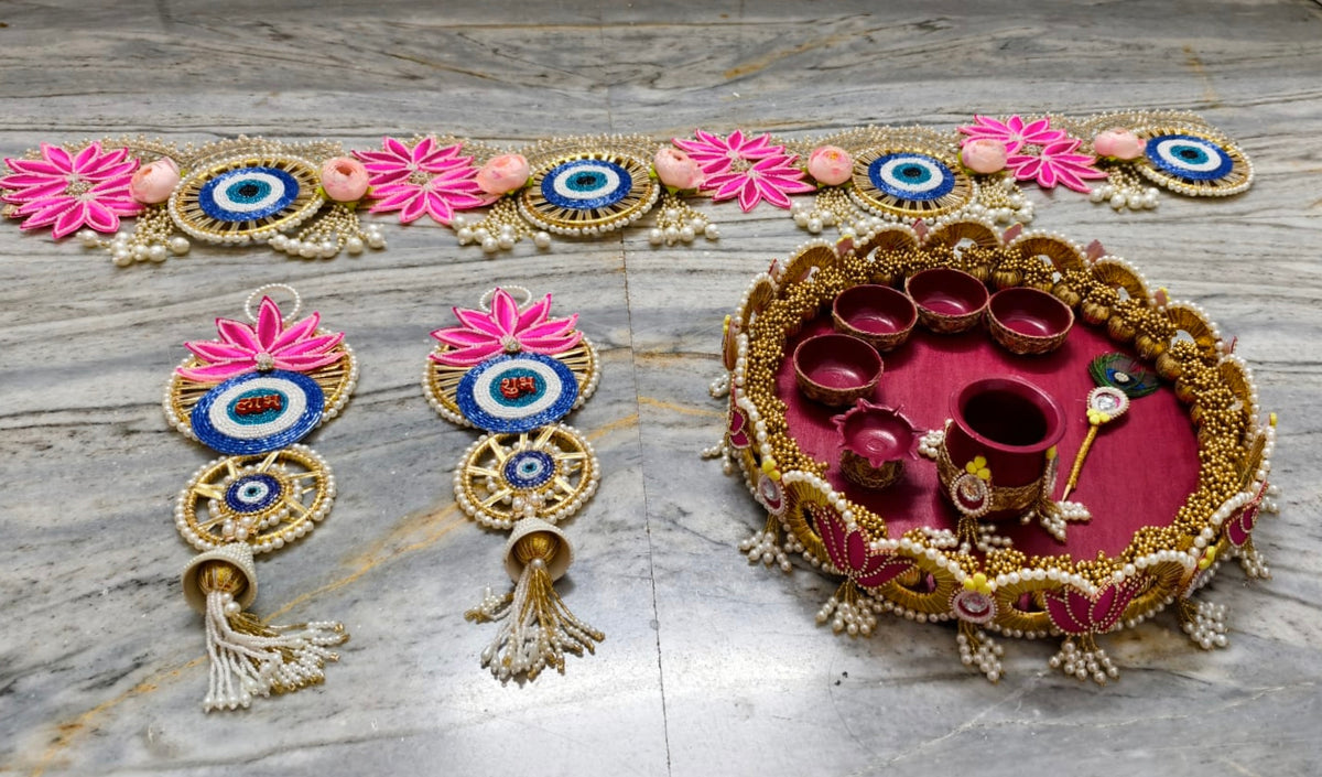 A decorative Diwali arrangement on a gray marble surface, featuring a long embroidered garland, two matching hanging ornaments with pink flowers and blue-and-white eye motifs, and a round gold tray lined with pink fabric and holding small red oil lamps.