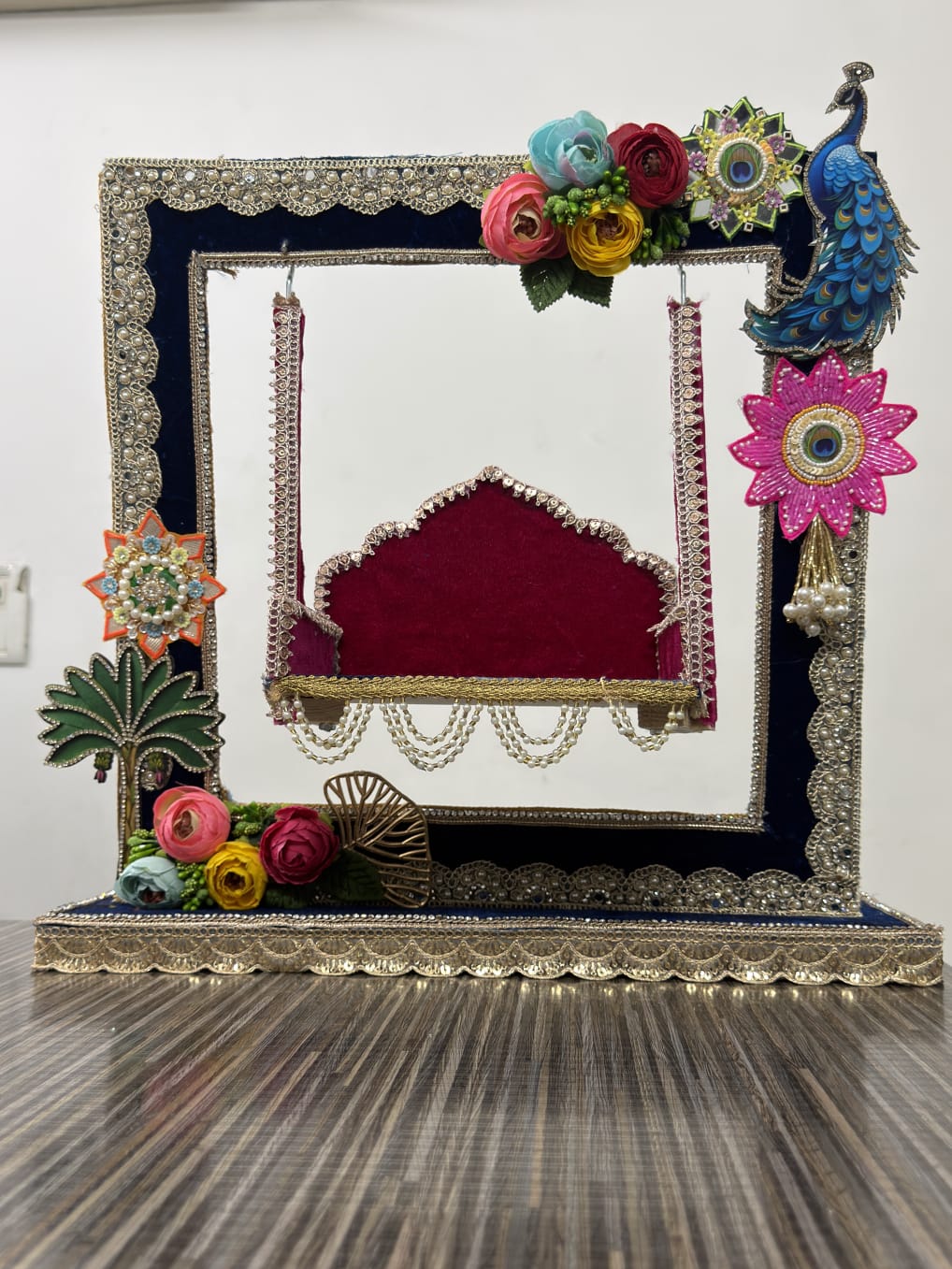 Handmade Krishna Jhula decorative swing with navy blue velvet frame, red velvet seat, pearl chains, colorful flowers and peacock motif