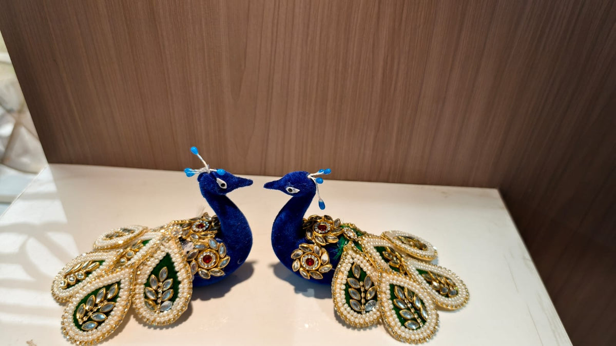 Two decorative peacock figurines with blue bodies and ornate gold, green, and white feather details, displayed on a white surface against a brown wood-grain background.
