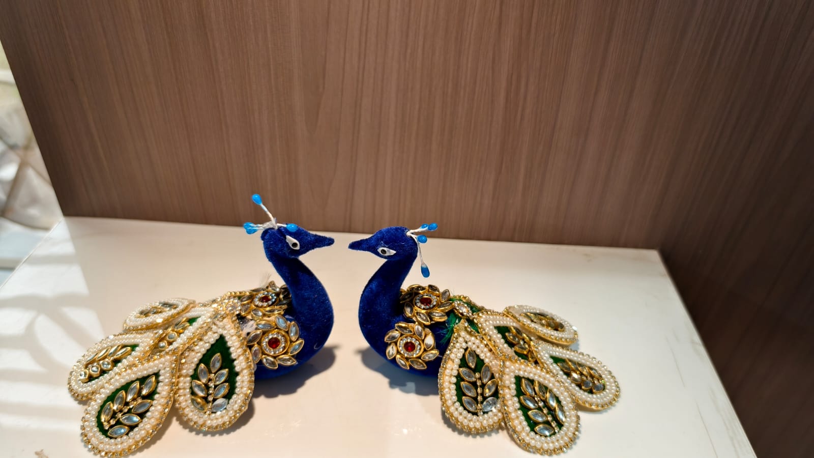 Two decorative peacock figurines with blue bodies and ornate gold, green, and white feather details, displayed on a white surface against a brown wood-grain background.
