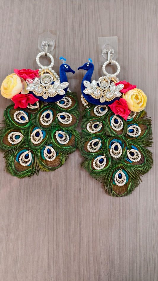 A pair of large, decorative peacock-themed earrings hanging on a light wood surface. Each earring features a blue peacock body, a green peacock tail with eye-like patterns, white faux pearls, silver rhinestone embellishments, and yellow and red fabric roses.